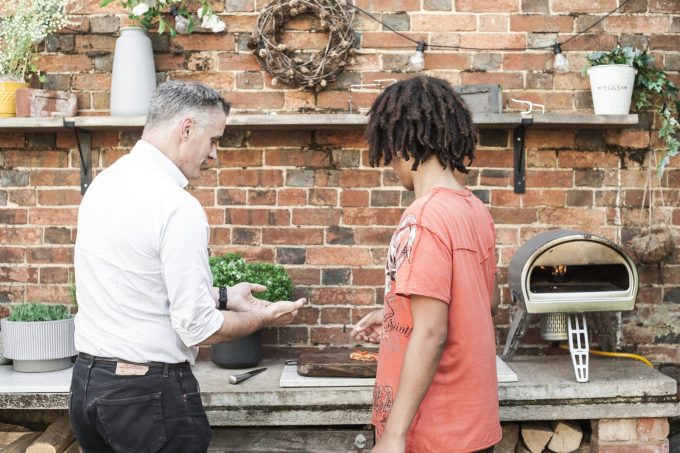 a man in a white shirt and black jeans talking to a teenage boy about pizza
